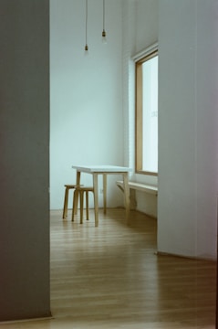 A minimalist room featuring a simple wooden table and stool set against a large window. The walls and floor are neutral in color, with exposed light bulbs hanging from the ceiling. The space appears open and uncluttered.