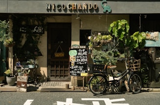 A quaint storefront with rustic charm features a sign reading 'Nico Chando' adorned with an elephant figure. The facade is decorated with lush greenery and hanging plants. In front of the entrance, a bicycle with a wicker basket is parked, and a chalkboard menu displays food items written in a foreign language. Potted plants of various shapes and sizes line the entrance, enhancing the inviting atmosphere.