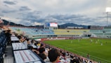 A lively soccer match is taking place in an outdoor stadium, viewed from the stands filled with spectators. The field is bright green, framed by the surrounding track, and players in jerseys are actively engaged in the game. The stadium features colorful seating areas in blue, yellow, and green, with a mountainous landscape visible in the background under a partly cloudy sky. A large digital scoreboard displays the match time and score.