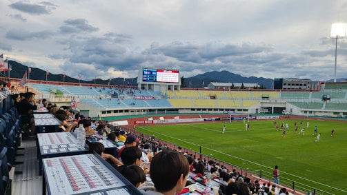A lively soccer match is taking place in an outdoor stadium, viewed from the stands filled with spectators. The field is bright green, framed by the surrounding track, and players in jerseys are actively engaged in the game. The stadium features colorful seating areas in blue, yellow, and green, with a mountainous landscape visible in the background under a partly cloudy sky. A large digital scoreboard displays the match time and score.