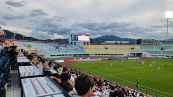 A lively soccer match is taking place in an outdoor stadium, viewed from the stands filled with spectators. The field is bright green, framed by the surrounding track, and players in jerseys are actively engaged in the game. The stadium features colorful seating areas in blue, yellow, and green, with a mountainous landscape visible in the background under a partly cloudy sky. A large digital scoreboard displays the match time and score.
