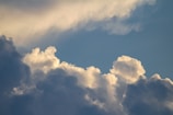 Cloud formations in a blue sky, with a mix of fluffy white clouds and darker, shadowed areas. Sunlight creates soft highlights on the edges of the clouds.