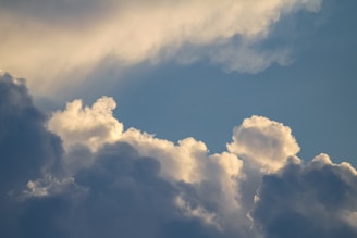 Cloud formations in a blue sky, with a mix of fluffy white clouds and darker, shadowed areas. Sunlight creates soft highlights on the edges of the clouds.