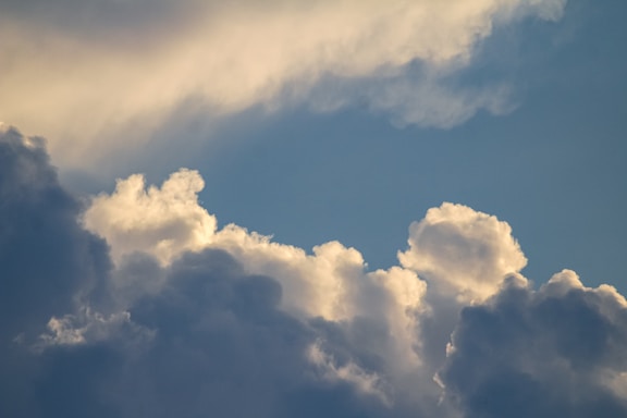 Cloud formations in a blue sky, with a mix of fluffy white clouds and darker, shadowed areas. Sunlight creates soft highlights on the edges of the clouds.