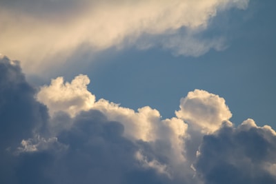 Cloud formations in a blue sky, with a mix of fluffy white clouds and darker, shadowed areas. Sunlight creates soft highlights on the edges of the clouds.