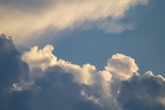 Cloud formations in a blue sky, with a mix of fluffy white clouds and darker, shadowed areas. Sunlight creates soft highlights on the edges of the clouds.