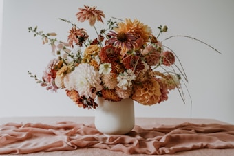 An elegant arrangement of mixed flowers in a variety of colors, including shades of pink, white, and orange, is placed in a simple white vase. The bouquet features a mix of bloom shapes and subtle greenery, set against a soft pink fabric laid on a flat surface.