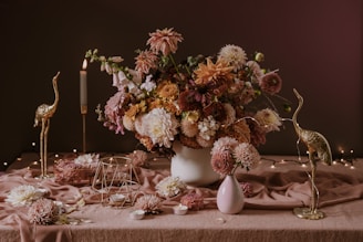 A richly arranged bouquet of flowers sits on a table draped with a soft, pinkish fabric. The flowers are in soft shades of pink, peach, and white, accompanied by green foliage. Flanked by gold-colored crane sculptures, the setting includes a lit candle, small candleholders, and decorative metal geometric shapes. The ambiance is enhanced by fairy lights in the background, creating a warm and elegant scene.