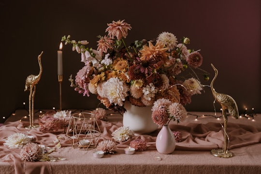 A richly arranged bouquet of flowers sits on a table draped with a soft, pinkish fabric. The flowers are in soft shades of pink, peach, and white, accompanied by green foliage. Flanked by gold-colored crane sculptures, the setting includes a lit candle, small candleholders, and decorative metal geometric shapes. The ambiance is enhanced by fairy lights in the background, creating a warm and elegant scene.