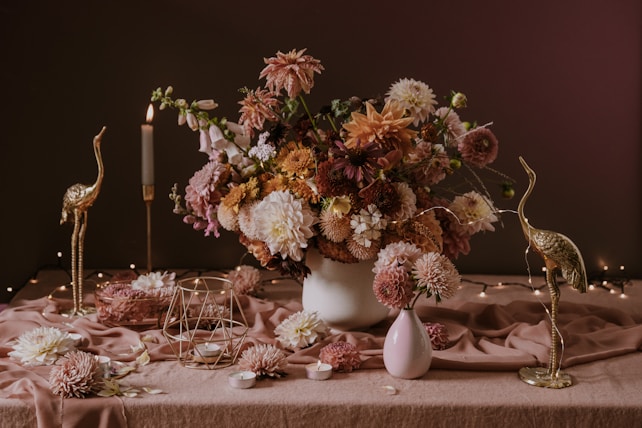 A richly arranged bouquet of flowers sits on a table draped with a soft, pinkish fabric. The flowers are in soft shades of pink, peach, and white, accompanied by green foliage. Flanked by gold-colored crane sculptures, the setting includes a lit candle, small candleholders, and decorative metal geometric shapes. The ambiance is enhanced by fairy lights in the background, creating a warm and elegant scene.