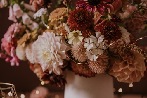A vibrant arrangement of diverse flowers in rich, earthy tones, including dahlias and chrysanthemums, is elegantly displayed in a soft-focus setting. The interplay of natural colors is complemented by delicate string lights interwoven among the floral arrangement.