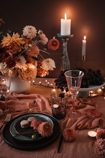 A beautifully arranged table setting with a centerpiece of vibrant flowers in shades of orange, pink, and cream. There are lit candles providing soft, warm lighting, along with elegant glassware and a dish featuring a couple of macarons. The table is adorned with a soft pink cloth and small flowers scattered around, contributing to the cozy, inviting atmosphere.