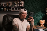 A person with tattoos and wearing a white t-shirt and cap is sitting in a chair, holding a smartphone. The room is decorated with a collection of action figures and a plush toy on the desk, along with a keyboard and mouse. The background wall is covered in artificial greenery.