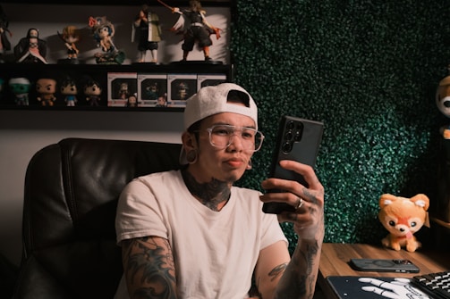 A person with tattoos and wearing a white t-shirt and cap is sitting in a chair, holding a smartphone. The room is decorated with a collection of action figures and a plush toy on the desk, along with a keyboard and mouse. The background wall is covered in artificial greenery.