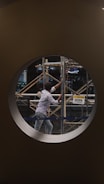 A person is working on scaffolding, visible through a circular window. The setting is a construction site, and there is a sign nearby reading 'Caution: Work in Progress'. The person is dressed in casual work attire, and the environment suggests an indoor area undergoing construction or renovation.