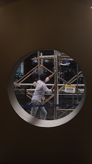 A person is working on scaffolding, visible through a circular window. The setting is a construction site, and there is a sign nearby reading 'Caution: Work in Progress'. The person is dressed in casual work attire, and the environment suggests an indoor area undergoing construction or renovation.