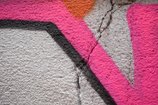 Bright pink and orange graffiti artwork on a rough, cracked concrete surface. The design featuring angular shapes is accented by a bold black line.
