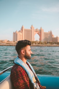 A person with a trimmed beard and wearing a life jacket sits in a boat. The background features the grand architecture of a large hotel or resort with an arch, possibly Atlantis The Palm in Dubai. The scene appears calm, with the gentle blue water surrounding the boat and a warm, late afternoon light illuminating the setting.