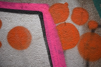 A textured wall features vibrant graffiti consisting of large orange circles and a prominent bold pink line bordered by black. The background is a rough stone surface, adding depth to the artwork.