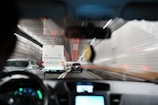 A view from inside a car driving through a tunnel. The scene shows the back of several vehicles, including a large white bus and a black car, all moving in the same direction. The tunnel's walls are lined with bright white tiles and red lights streak along the ceiling, creating a sense of speed and motion.