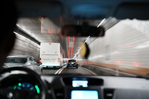A view from inside a car driving through a tunnel. The scene shows the back of several vehicles, including a large white bus and a black car, all moving in the same direction. The tunnel's walls are lined with bright white tiles and red lights streak along the ceiling, creating a sense of speed and motion.