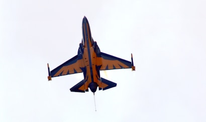 A fighter jet is captured flying upwards with its afterburners glowing. The aircraft has a striking design with bold patterns in blue and orange on its body and wings against a light, cloudy sky.