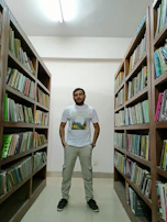 A person stands confidently in the middle of a library aisle, surrounded by shelves packed with books. The individual is wearing a casual outfit consisting of a t-shirt, cargo pants, and sneakers. The library has a well-lit, organized appearance with wooden shelves and a light-colored ceiling.