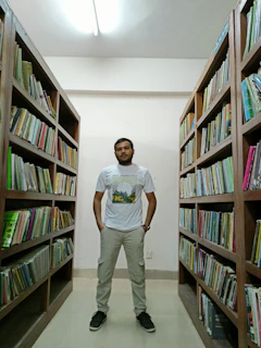 A person stands confidently in the middle of a library aisle, surrounded by shelves packed with books. The individual is wearing a casual outfit consisting of a t-shirt, cargo pants, and sneakers. The library has a well-lit, organized appearance with wooden shelves and a light-colored ceiling.