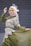 A person wearing an elegant green dress with intricate white lace patterns, accessorized with a light-colored headscarf and a decorative fascinator. The individual stands gracefully against a textured stone wall background, striking a poised and sophisticated pose.