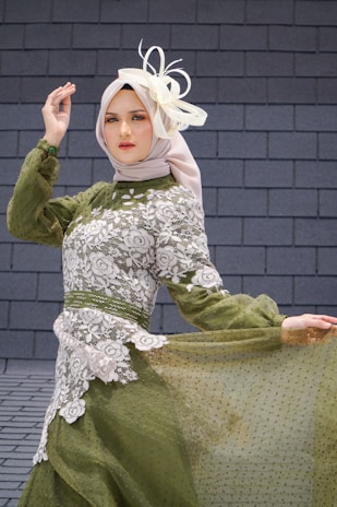 A person wearing an elegant green dress with intricate white lace patterns, accessorized with a light-colored headscarf and a decorative fascinator. The individual stands gracefully against a textured stone wall background, striking a poised and sophisticated pose.