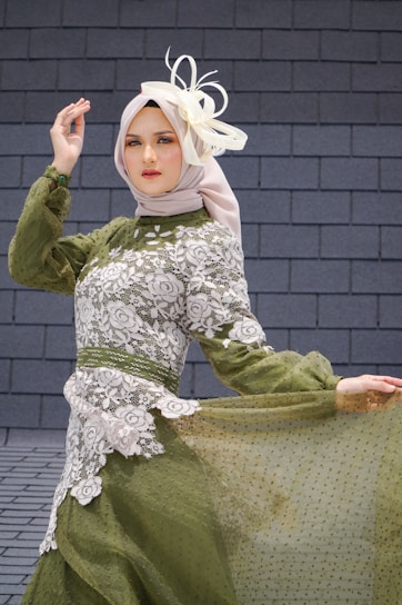 A person wearing an elegant green dress with intricate white lace patterns, accessorized with a light-colored headscarf and a decorative fascinator. The individual stands gracefully against a textured stone wall background, striking a poised and sophisticated pose.