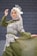 A person wearing an elegant green dress with intricate white lace patterns, accessorized with a light-colored headscarf and a decorative fascinator. The individual stands gracefully against a textured stone wall background, striking a poised and sophisticated pose.