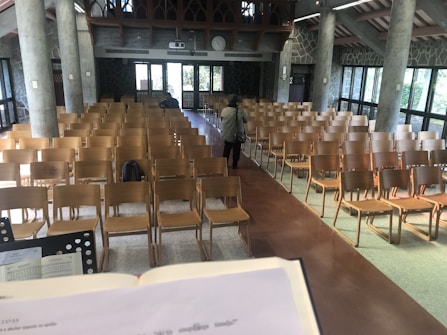 A large room with rows of wooden chairs facing toward the front, where a person is seen walking away. A podium or lectern with an open book can be seen in the foreground. The walls are made of stone with large windows letting in natural light. The room is adorned with arched wooden details at the top and there are concrete pillars evenly spaced along the walls.