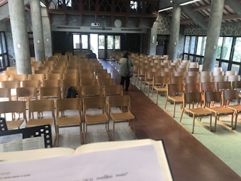 A large room with rows of wooden chairs facing toward the front, where a person is seen walking away. A podium or lectern with an open book can be seen in the foreground. The walls are made of stone with large windows letting in natural light. The room is adorned with arched wooden details at the top and there are concrete pillars evenly spaced along the walls.