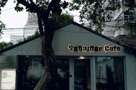 A quaint caf&eacute; with a rustic exterior nestled among trees. The sign reads 'Sabaidee Cafe' in illuminated letters. A tree partially obscures the front, adding a cozy and natural feel. Urban buildings are visible in the background.