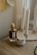 A perfume bottle with a decorative animal-shaped cap is placed on a wooden surface next to a small jar with a metal lid. In the background, a stand holds several tall, white, unlit candles. The setting appears to be dimly lit, creating a cozy and intimate atmosphere.