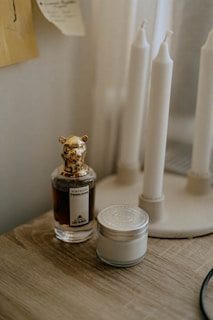 A perfume bottle with a decorative animal-shaped cap is placed on a wooden surface next to a small jar with a metal lid. In the background, a stand holds several tall, white, unlit candles. The setting appears to be dimly lit, creating a cozy and intimate atmosphere.