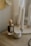 A perfume bottle with a decorative animal-shaped cap is placed on a wooden surface next to a small jar with a metal lid. In the background, a stand holds several tall, white, unlit candles. The setting appears to be dimly lit, creating a cozy and intimate atmosphere.