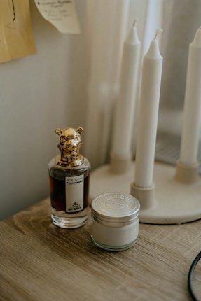 A perfume bottle with a decorative animal-shaped cap is placed on a wooden surface next to a small jar with a metal lid. In the background, a stand holds several tall, white, unlit candles. The setting appears to be dimly lit, creating a cozy and intimate atmosphere.