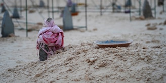 A simple stone marker draped with a pink cloth stands at the head of a sandy grave mound. Next to the mound, a shallow dish sits on the ground. The background shows a few indistinct grave markers and fencing.