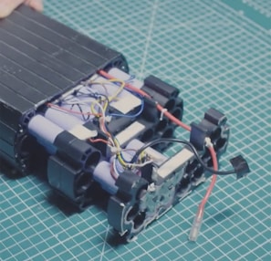 A partially assembled battery pack with visible cylindrical cells and multicolored electrical wires on a cutting mat with a grid pattern. The components appear organized, with several blue, red, black, and yellow wires intertwined among the battery cells.