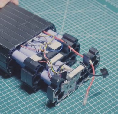 A partially assembled battery pack with visible cylindrical cells and multicolored electrical wires on a cutting mat with a grid pattern. The components appear organized, with several blue, red, black, and yellow wires intertwined among the battery cells.