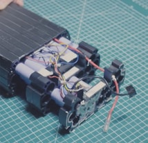 A partially assembled battery pack with visible cylindrical cells and multicolored electrical wires on a cutting mat with a grid pattern. The components appear organized, with several blue, red, black, and yellow wires intertwined among the battery cells.