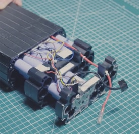 A partially assembled battery pack with visible cylindrical cells and multicolored electrical wires on a cutting mat with a grid pattern. The components appear organized, with several blue, red, black, and yellow wires intertwined among the battery cells.