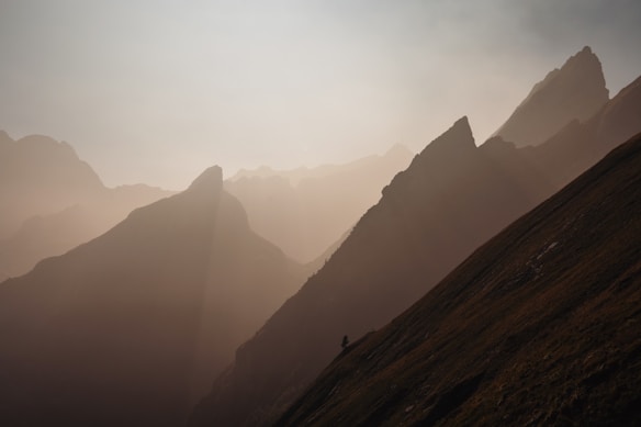 The image features a breathtaking view of mountain peaks partially shrouded in mist, with a soft, warm light casting gentle shadows. The slopes are steep, and the scene conveys a sense of serenity and vastness.