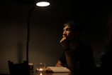 A person is sitting at a dimly lit table, appearing deep in thought. A single overhead lamp casts a focused light on the individual and on an open book in front of them. A small candle on the table provides additional soft illumination, creating a contemplative and intimate atmosphere.