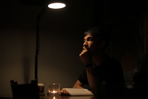 A person is sitting at a dimly lit table, appearing deep in thought. A single overhead lamp casts a focused light on the individual and on an open book in front of them. A small candle on the table provides additional soft illumination, creating a contemplative and intimate atmosphere.