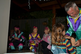 A group of people are gathered in a cozy indoor setting, wearing colorful retro-style tracksuits. There are visible decorations, including hanging lights and plants. The mood appears lively and social, with individuals engaged in conversation and interaction.