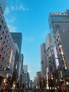 A bustling city street with tall modern buildings, featuring numerous signs and advertisements. The sky is clear and blue, creating a vibrant cityscape. People are walking along the sides, indicating a lively urban atmosphere.