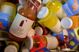 A collection of colorful bottled juices with labels, featuring a prominent brown bottle labeled 'Sapje' in the foreground. The labels are written in Dutch and include phrases related to health. The bottles vary in color with caps in different hues.